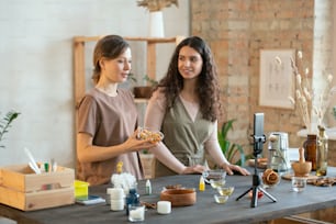 Hands of two young women cutting hard soap mass into cubes on boards while standing by table with ingredients for making cosmetic products