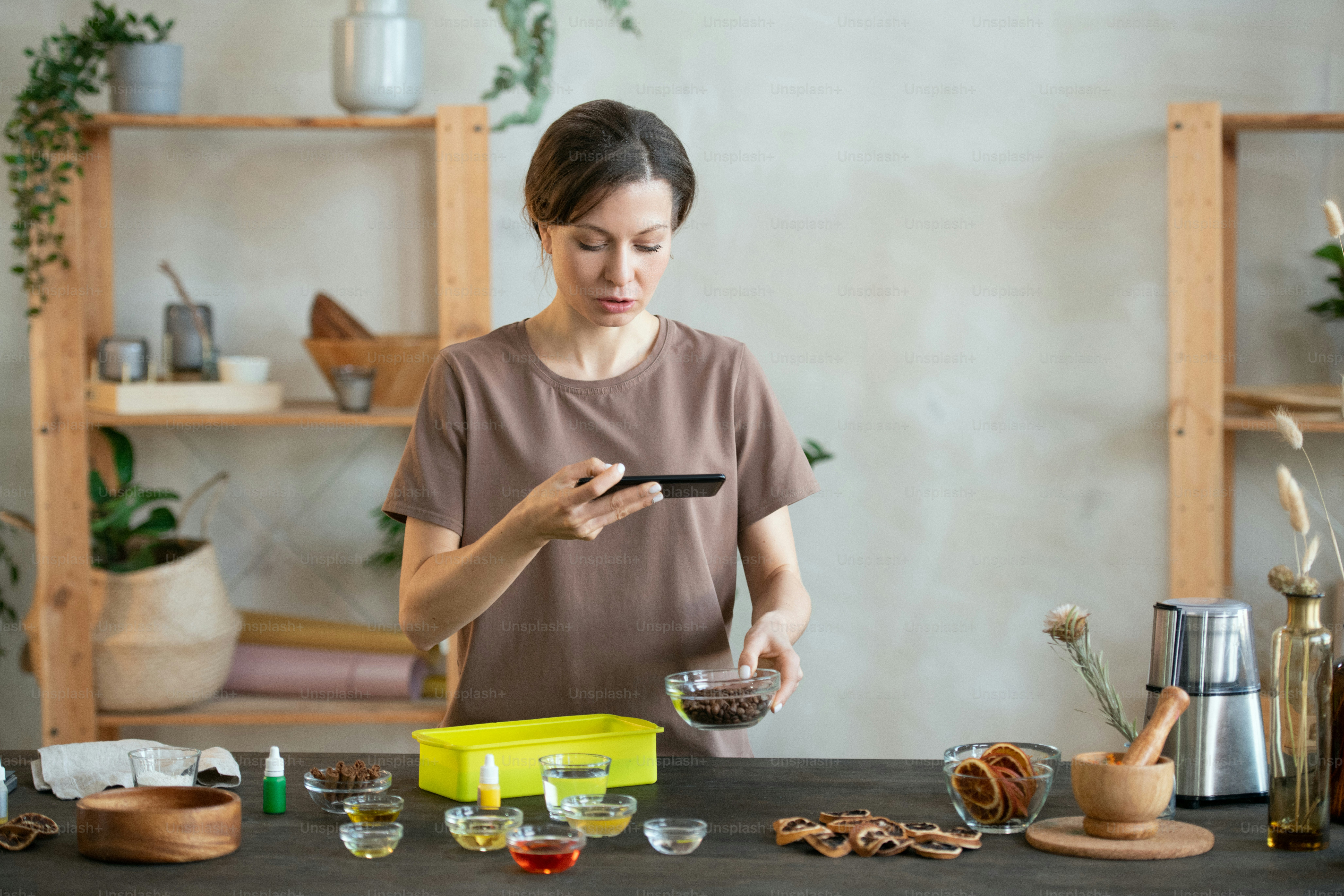 Jeune femme avec un smartphone filmant une vidéo debout près de la table et touchant un petit bol contenant de l’huile d’essence pour savon fait à la main
