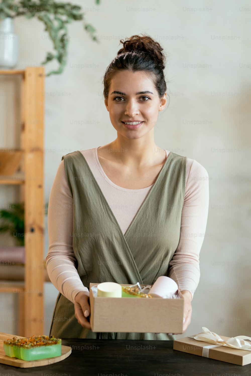 Young cheerful female in smart casualwear holding cardboard giftbox with handmade packed present while standing by table in front of camera