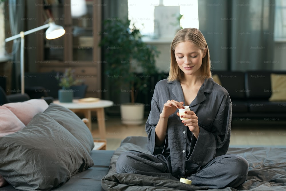 Young blond pretty female in grey pajamas applying natural handmade cosmetic product on her face while sitting on bed in the morning
