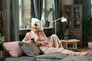 Young gorgeous woman in silk pajamas, towel on head and golden cosmetic mask on face relaxing on bed and scrolling in smartphone