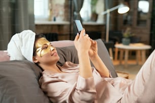 Young beautiful woman in silk pajamas, towel on head and golden cosmetic mask on face relaxing on bed and making selfie