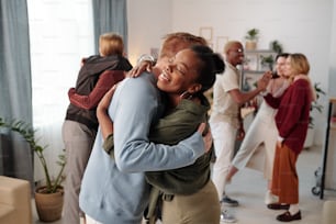 Happy young African woman in smart casualwear giving hug to guy in blue pullover in front of camera against their friends embracing and talking