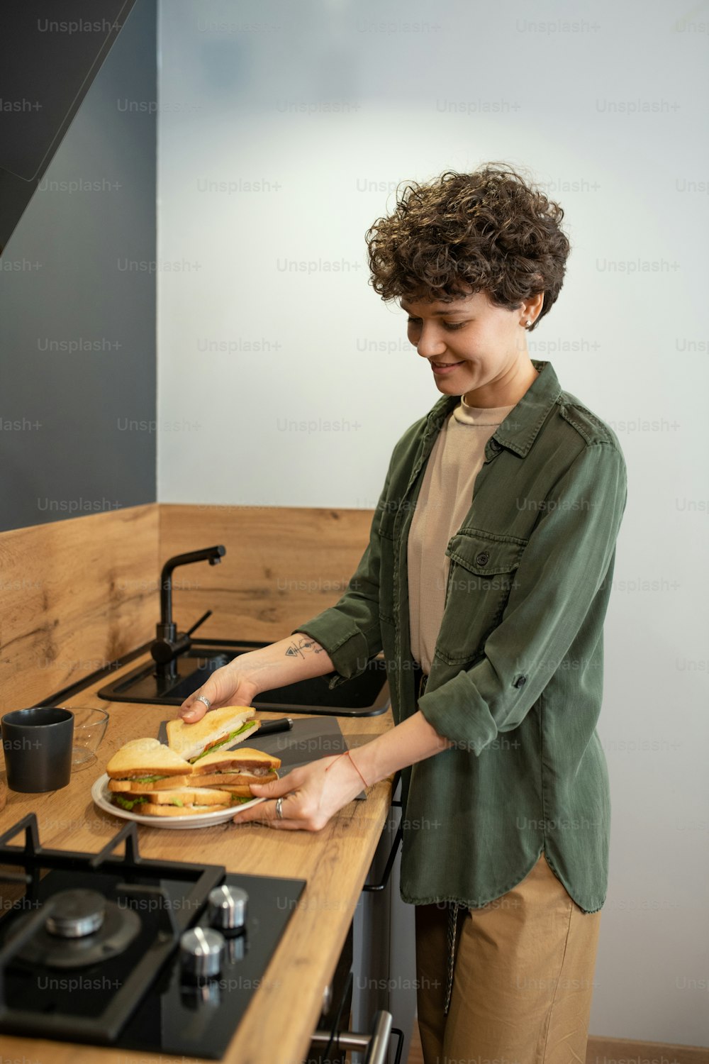 Happy young brunette female in casualwear putting homemade sandwiches on plate while standing between sink and stove in the kitchen