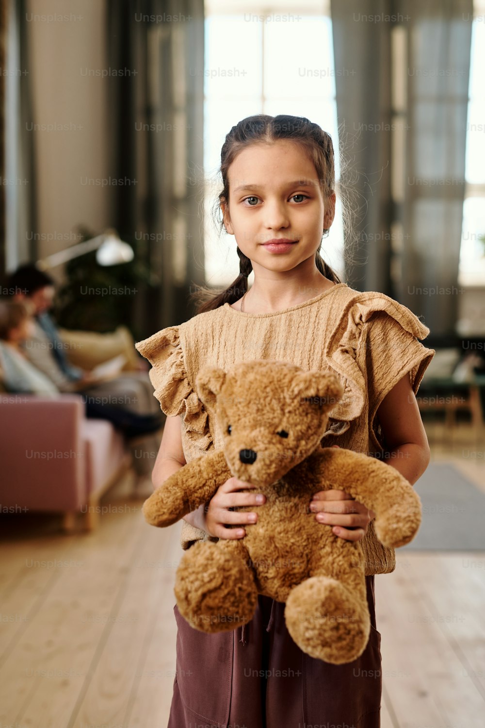 Little pretty girl with soft brown teddybear looking at you while standing in front of camera against her family sitting on couch in living-room