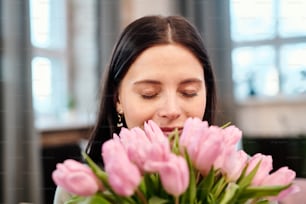 Jeune femme brune heureuse gardant les yeux fermés tout en tenant un bouquet de tulipes roses fraîches par le nez et en appréciant leur parfum