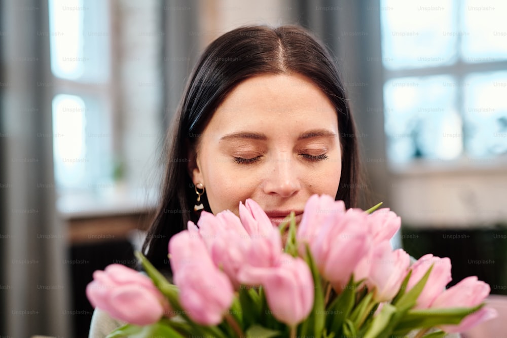 Happy young brunette female keeping her eyes closed while holding bunch of fresh pink tulips by her nose and enjoying their fragrance