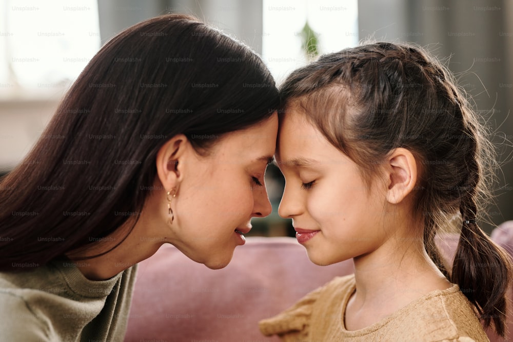 Side view of young brunette woman and her cute little daughter in casualwear touching by foreheads while standing in front of camera