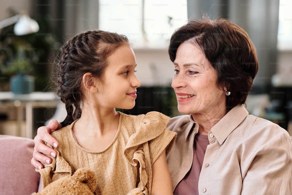 Affectionate mature brunette woman embracing her cute little granddaughter while both looking at one another with smiles