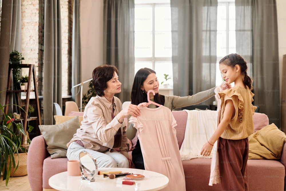 Two elegant women sitting on couch in living-room and showing new dresses to adorable little girl after buying them in online shop