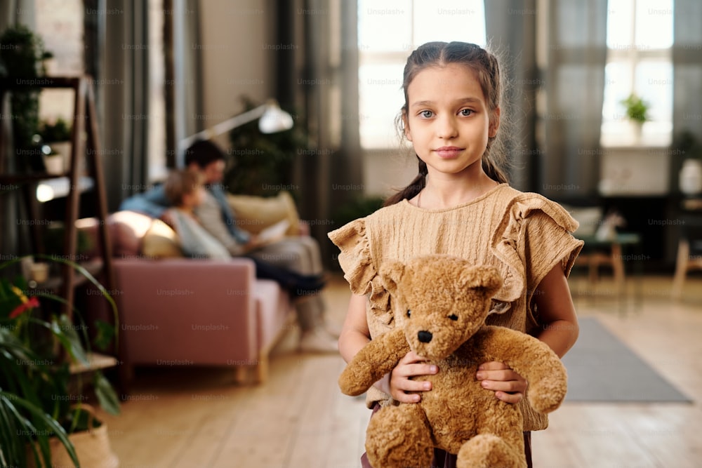 Menina bonita com ursinho de pelúcia em pé na sala de estar contra sua família