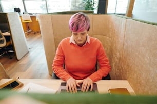 Contemporary businesswoman or office manager in casualwear looking at laptop display while sitting in armchair by desk and networking
