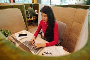 Contemporary Asian businesswoman or office manager in smart casualwear looking at laptop display while sitting by desk and networking