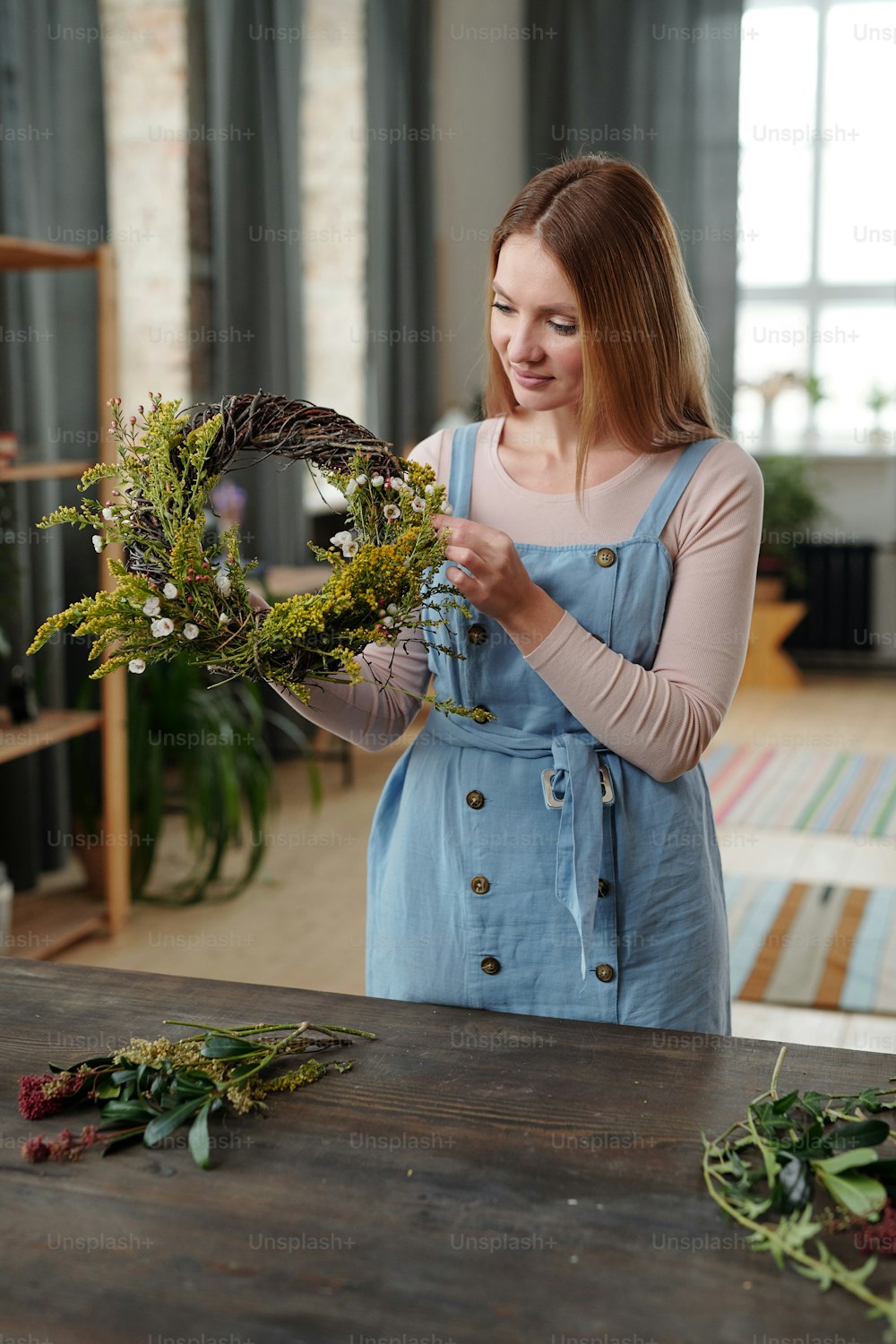 Happy young blond female in casualwear standing by wooden table in home environment and preparing Easter wreath for holiday