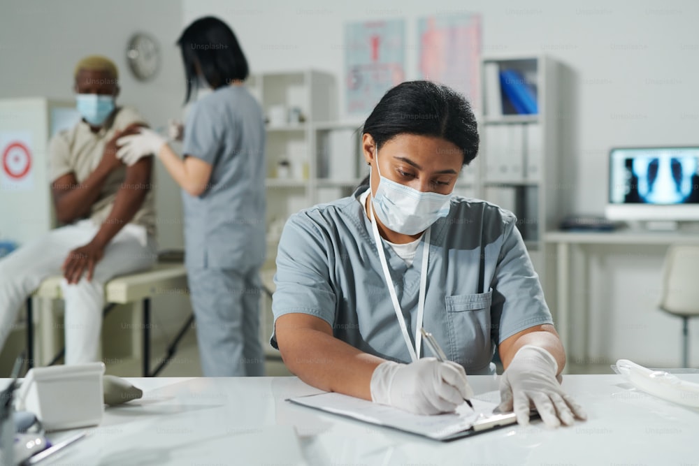 Young mixed-race clinician in protective workwear sitting by desk and filling in document while her colleague vaccinating male patient