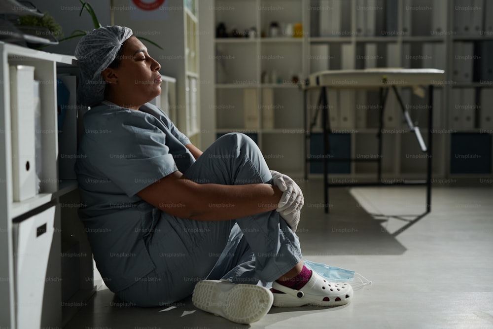 Seitenansicht einer jungen, müden, gemischtrassigen Klinikerin, die die Augen geschlossen hält, während sie auf dem Boden sitzt und sich nach einem harten Arbeitstag ausruht