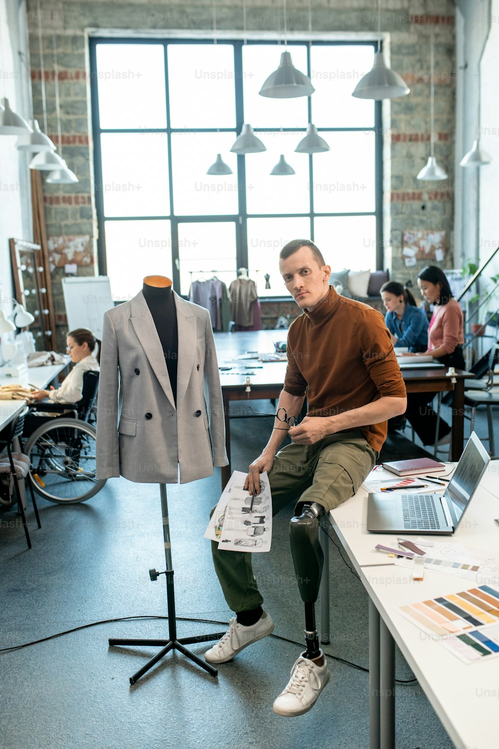 Un jeune créateur de mode handicapé avec des croquis assis sur la table devant la caméra contre des collègues féminines occupées dans un grand atelier
