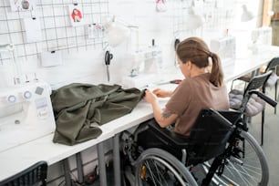 Junge behinderte Frau in Freizeitkleidung, die im Rollstuhl am Arbeitsplatz in einer großen zeitgenössischen Werkstatt sitzt und Kleidung näht