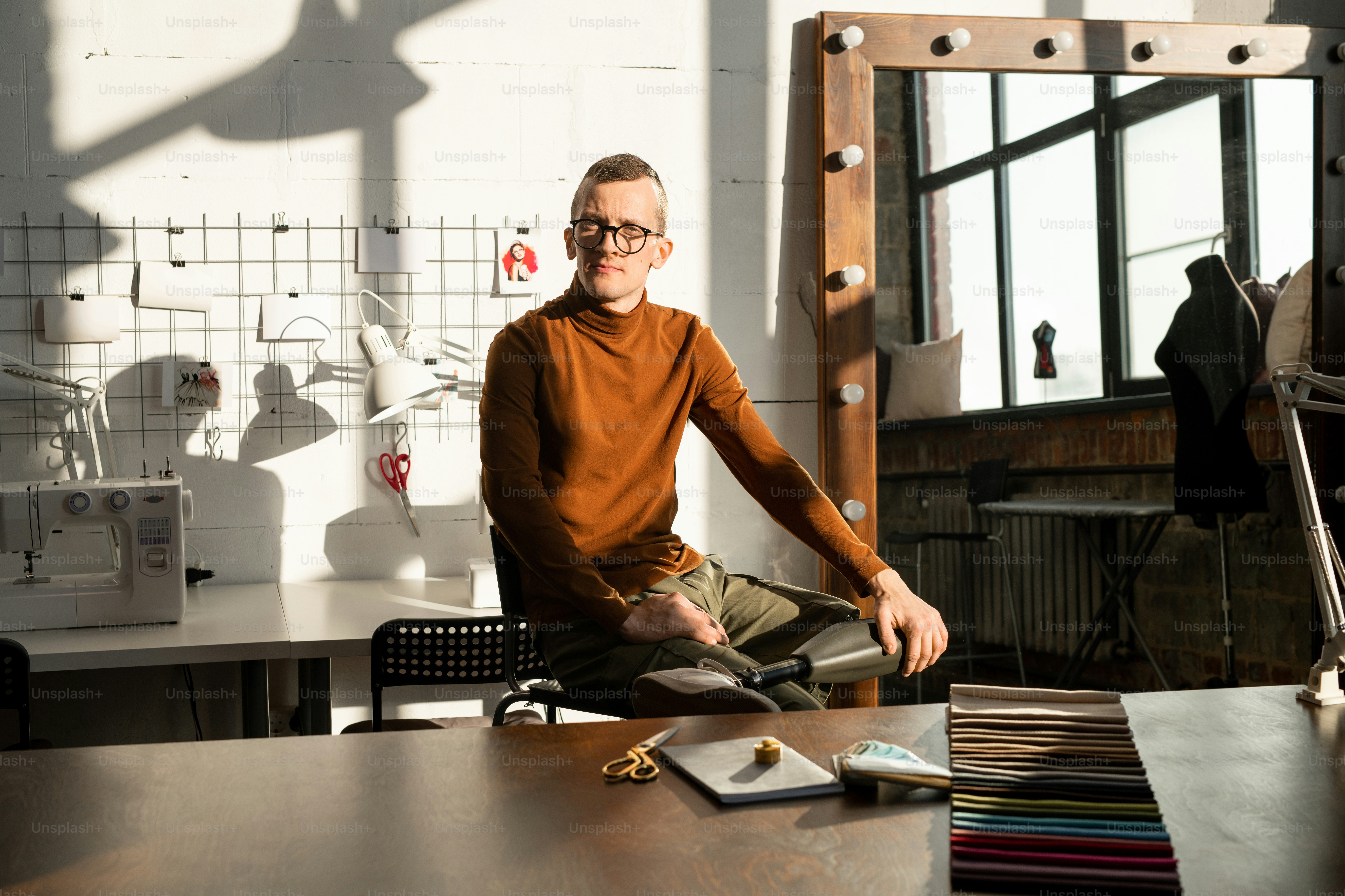 Young serious male tailor with prosthesis instead of left leg sitting by workplace in front of camera inside large contemporary workshop