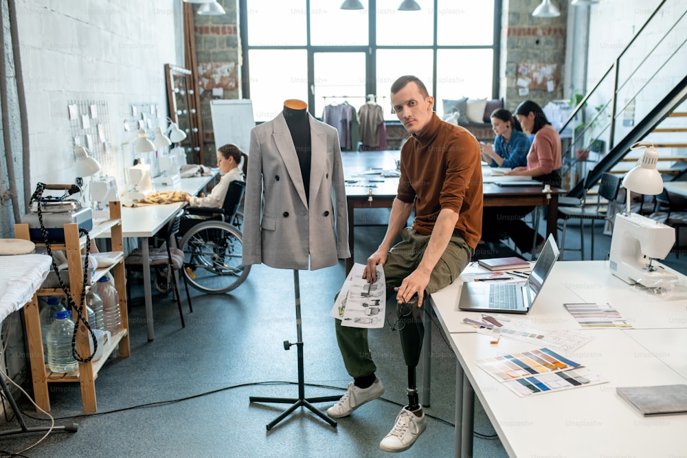 Young serious fashion designer or tailor with prosthetic leg sitting on table by mannequin against busy female colleagues inside large workshop