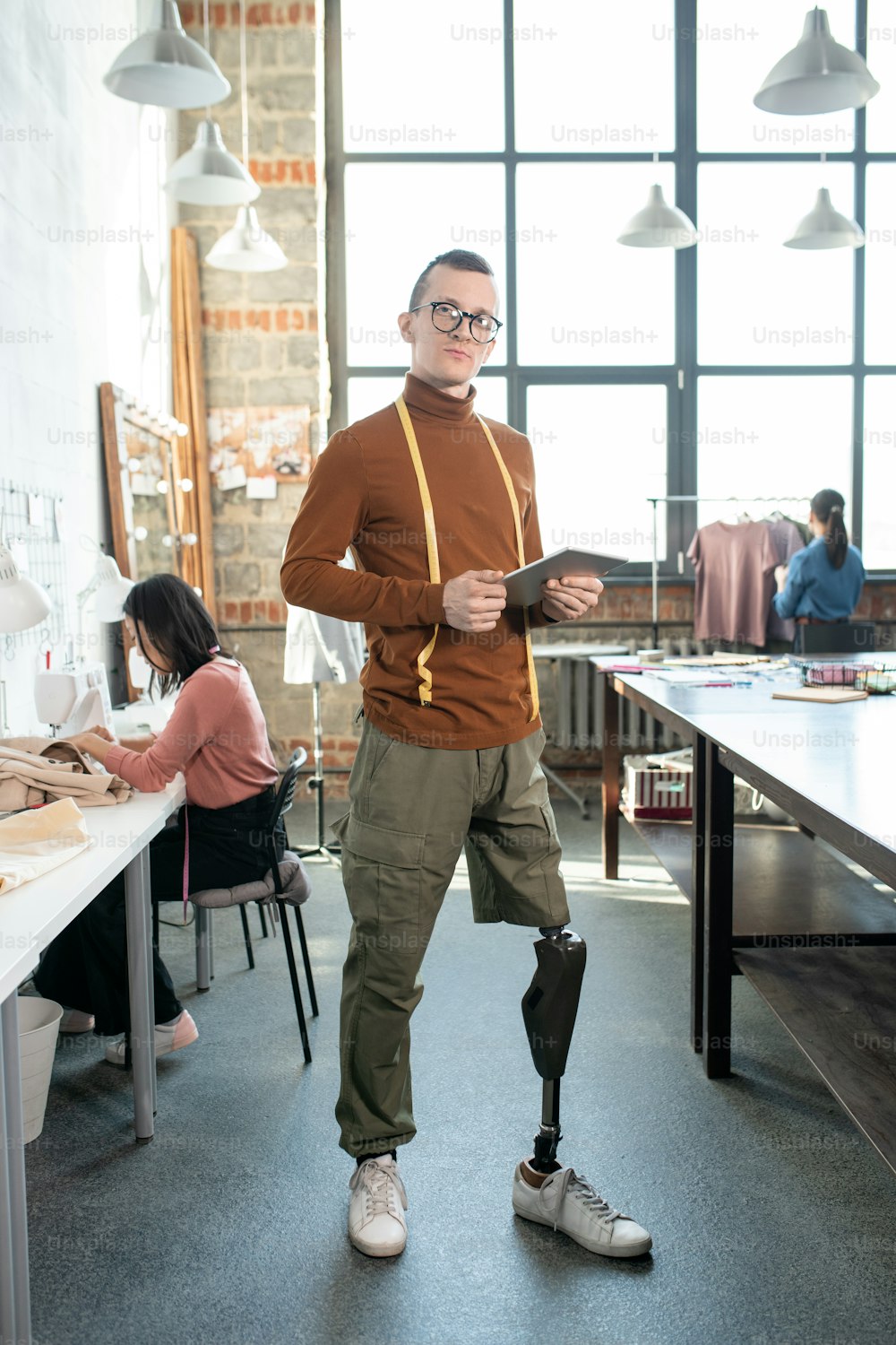 Young fashion designer or tailor with prosthetic leg using tablet by workplace against female colleagues inside large workshop