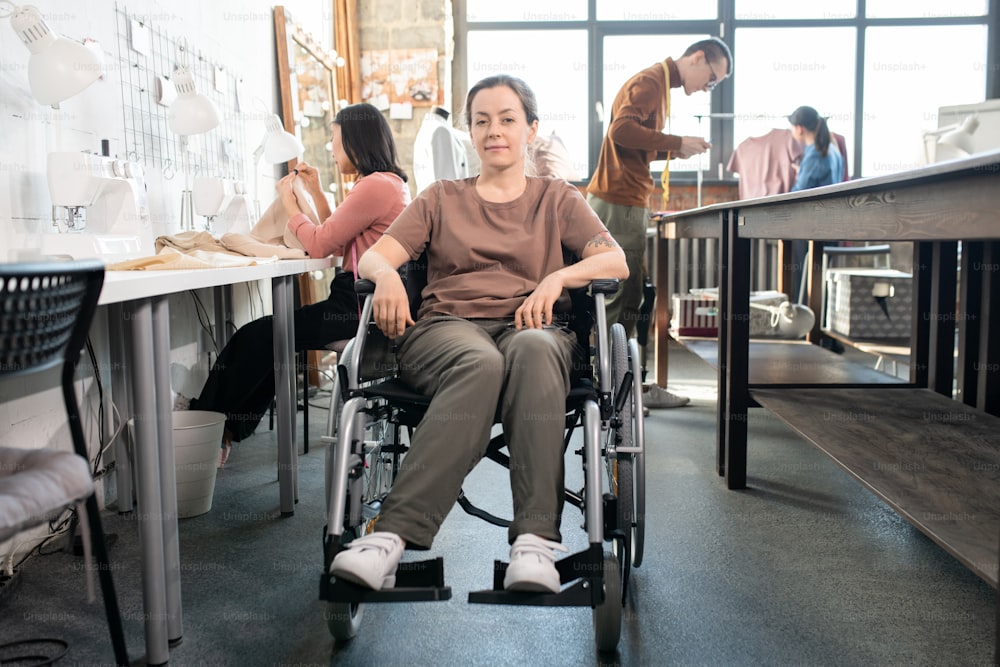 Young disable female in casualwear sitting in wheelchair by workplace against group of colleagues working over new fashion collection