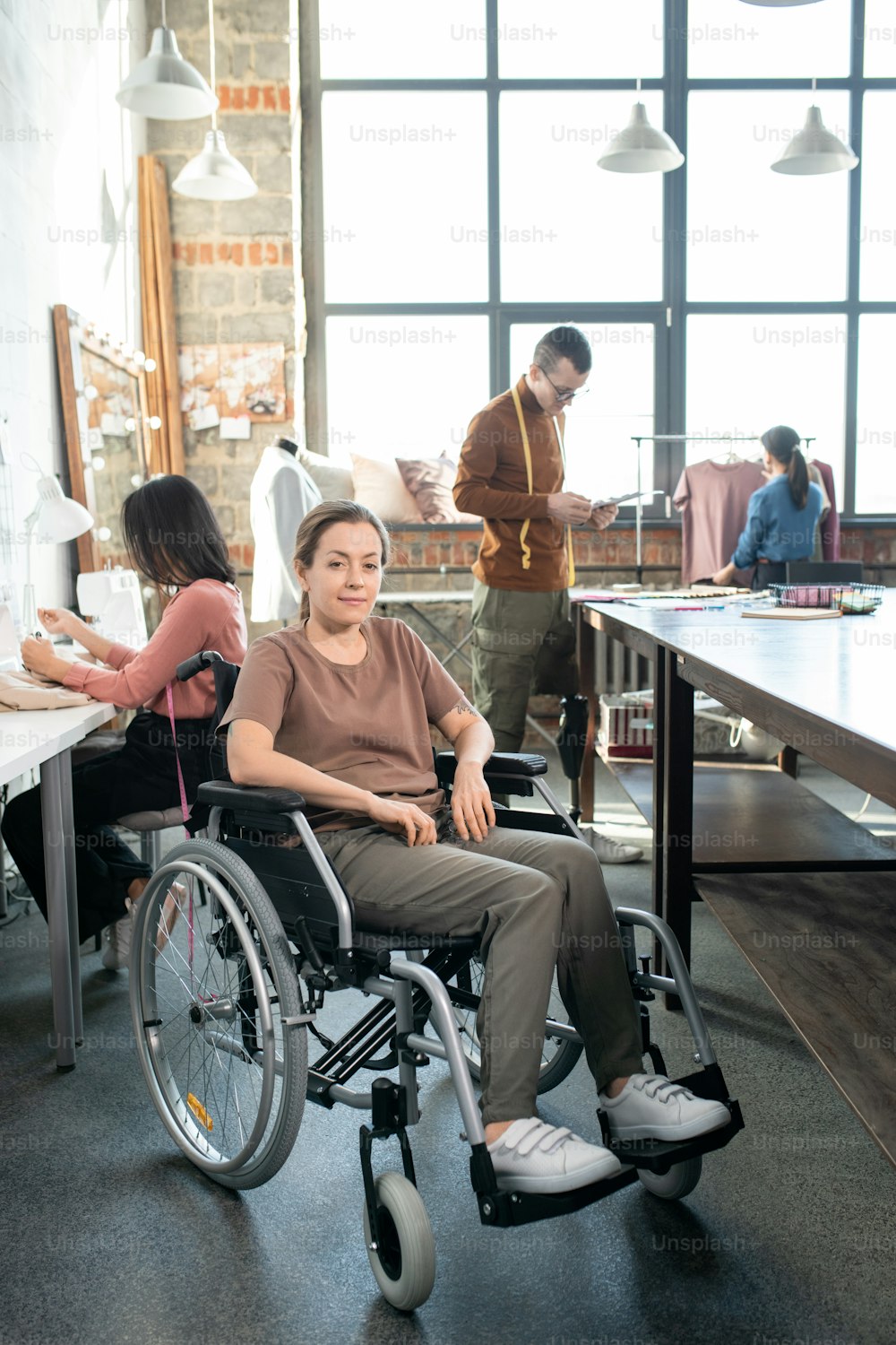Young disable seamstress in casualwear sitting in wheelchair in front of camera against colleagues working over new fashion collection
