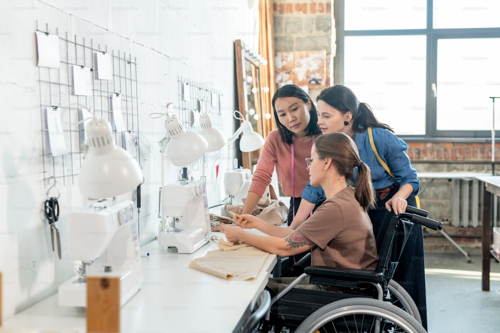 Two young female fashion designers consulting with disable seamtsress in wheelchair during discussion of the main points of new collection