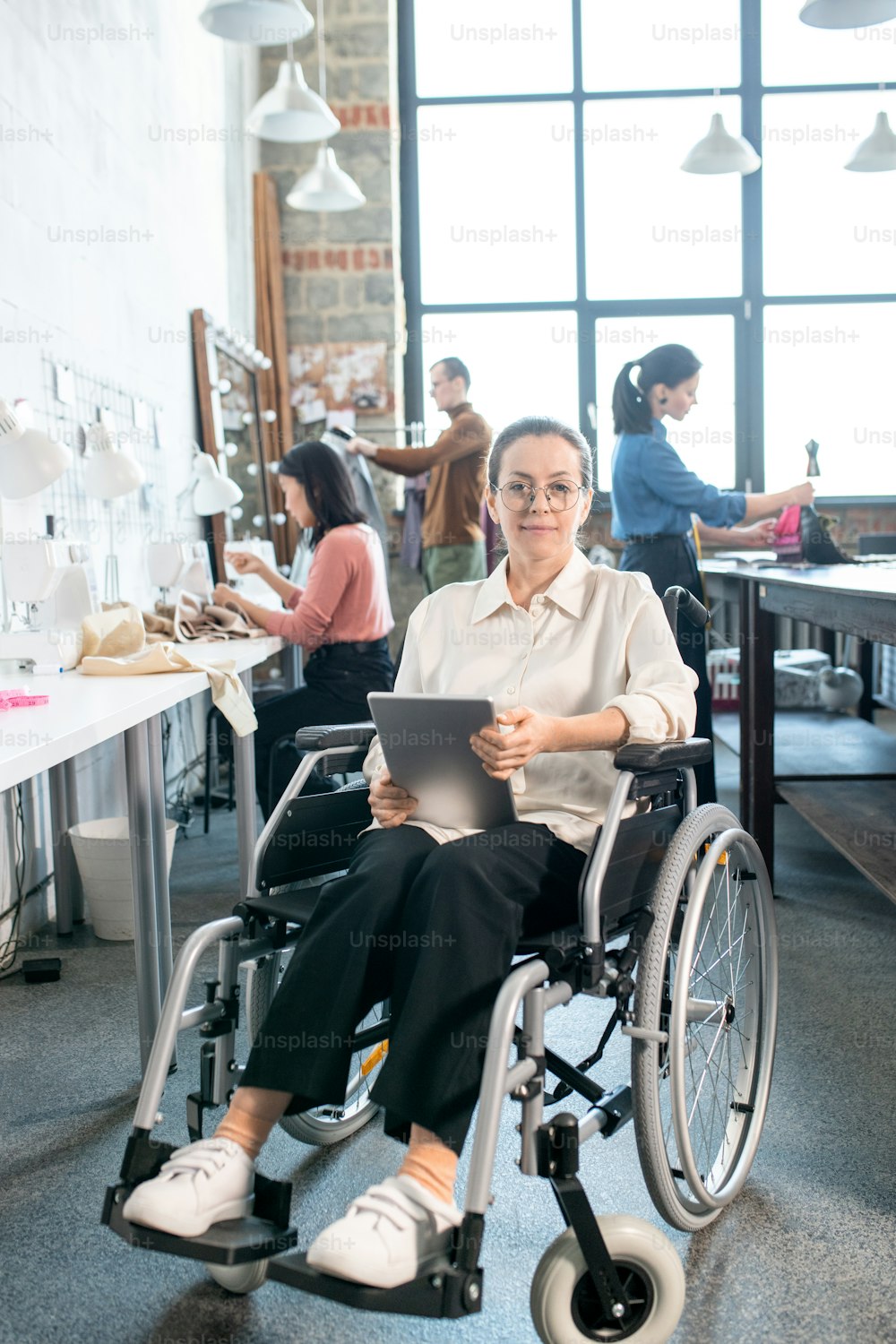 Young smiling seamstress with tablet sitting in wheelchair against group of colleagues working over new fashion collection in large workshop