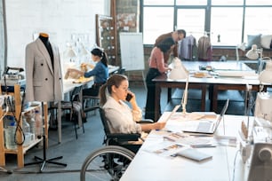 Young creative designer in wheelchair sitting by desk, looking through papers with new sketches and consulting one of clients on the phone