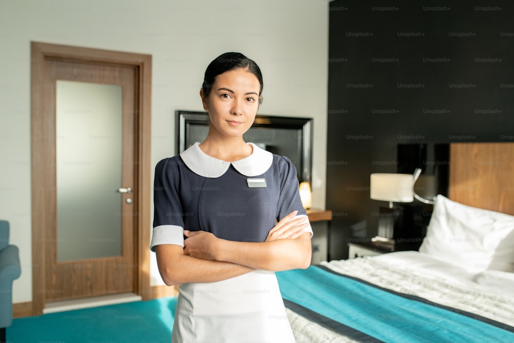 Young beautiful brunette chambermaid in uniform keeping her arms crossed by chest while standing against done bed in hotel room