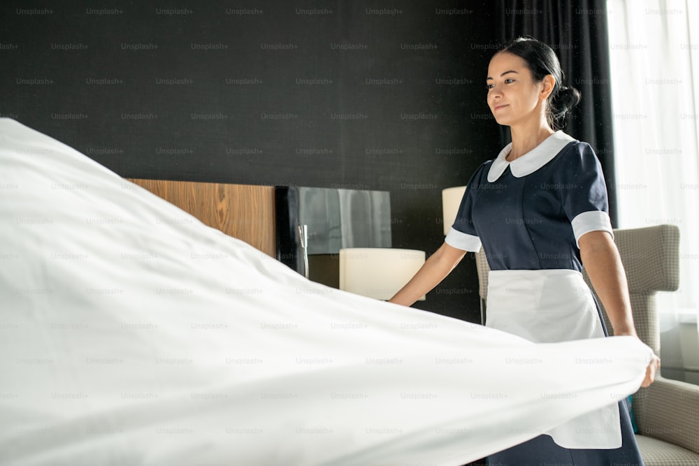 Young smiling pretty chambermaid putting white clean linen while changing bed in hotel room during preparation for new guests
