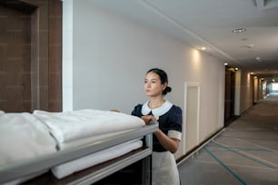 Giovane cameriera ai piani bruna o lavoratore dell'hotel che spinge il carrello con asciugamani puliti piegati e altre cose mentre si muove lungo il corridoio