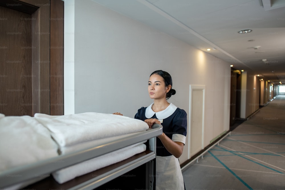 Young pretty brunette chambermaid or hotel worker pushing cart with folded clean towels and other stuff while moving along corridor