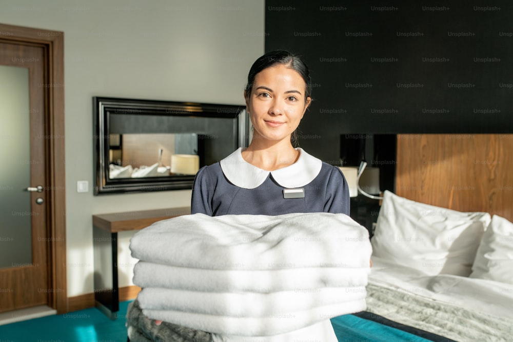 Personal de servicio de habitaciones feliz sosteniendo pila de sábanas blancas limpias en el dormitorio