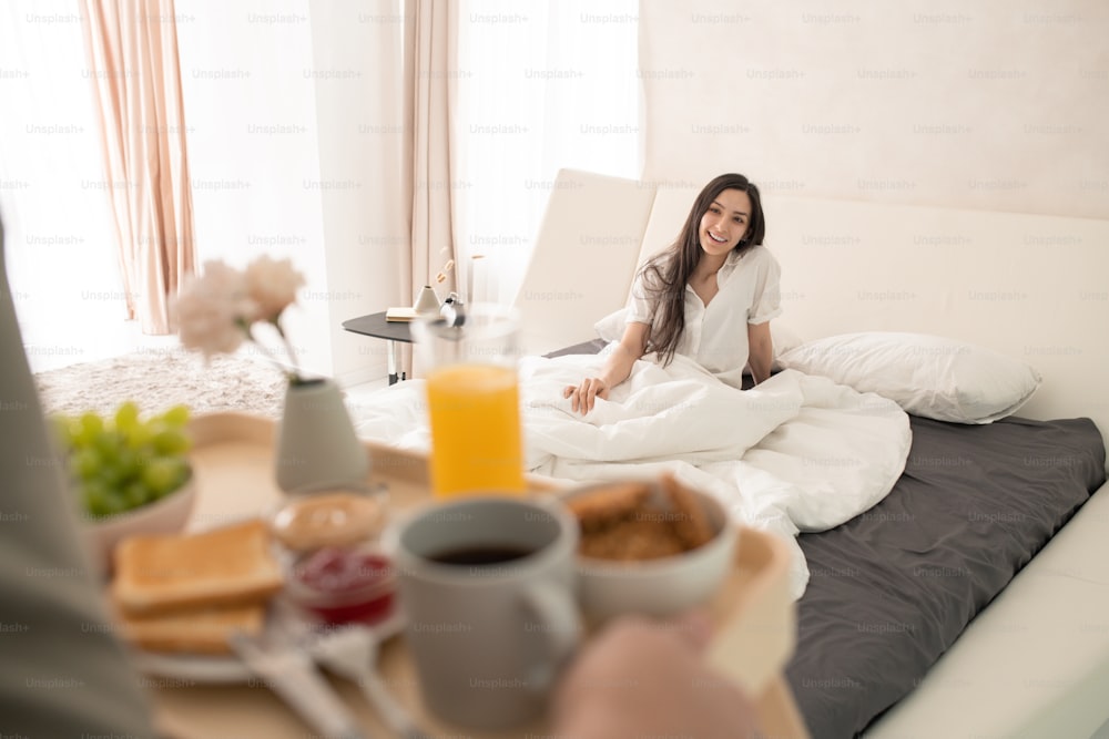 Happy young brunette female sitting under white blanket on large double bed and looking at her husband carrying tray with breakfast