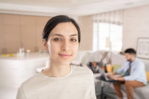 Young gorgeous woman in white t-shirt looking at you while standing in front of camera in living-room against her husband using laptop
