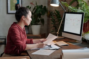 Young serious female architect in casualwear looking at sketch on paper while sitting by table in front of computer screen with picture of house