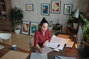 Young serious female architect with pencil drawing sketch of new construction while sitting by table in front of computer monitor