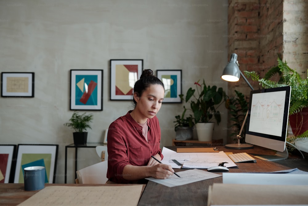 Young serious female architect with pencil pointing at sketch on paper while sitting by table in front of computer screen with picture of house