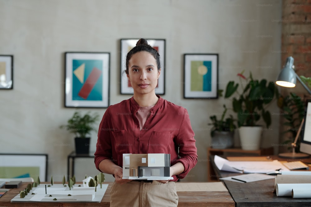 Contemporary young female architect or designer in casualwear holding paper model of new house while standing in office environment