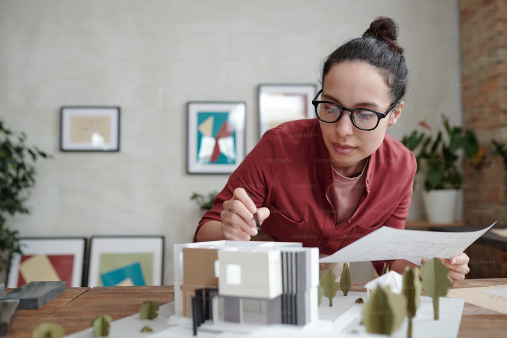 Young elegant female architect with paper and pen bending over model of new house and yard and pointing at roof while working over project