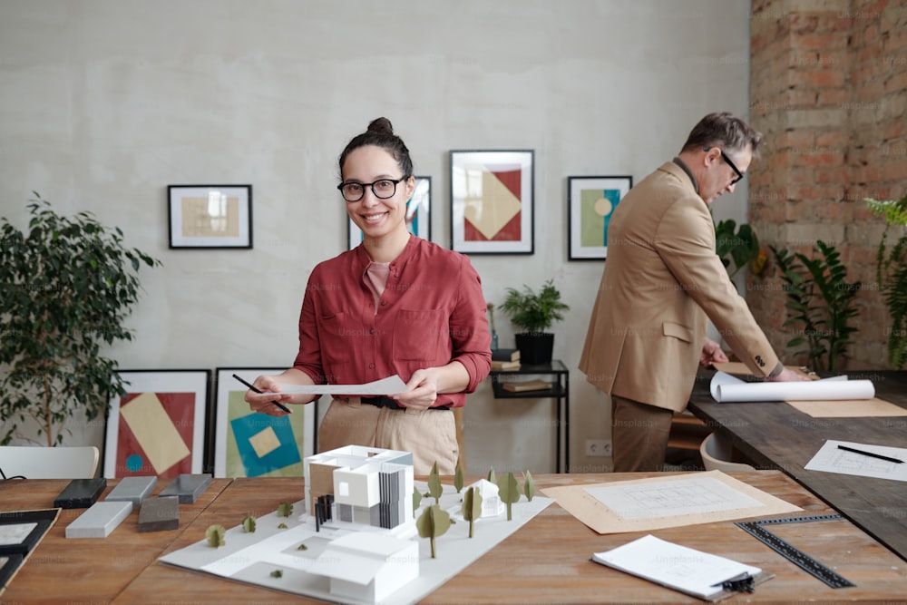Young smiling female architect in casualwear and eyeglasses standing by table with model of new house and yard against mature male colleague
