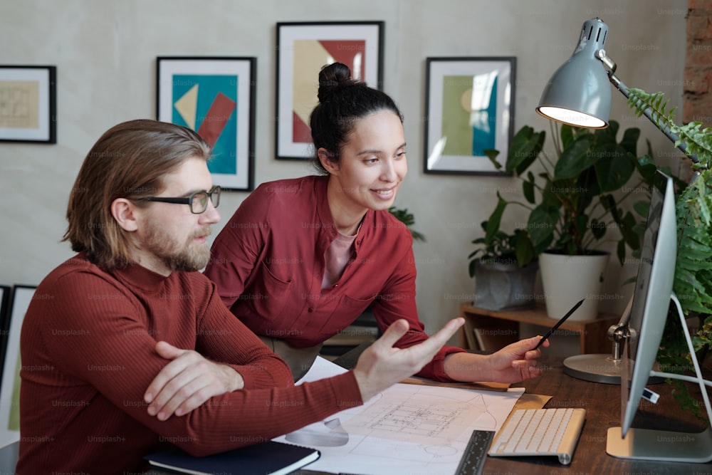 Positive young interracial architects in burgundy outfits looking at computer monitor and discussing online sketch in office