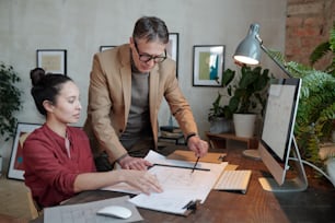 Young mixed race engineer sitting at desk with lamp and computer and discussing blueprint with boss in contemporary office