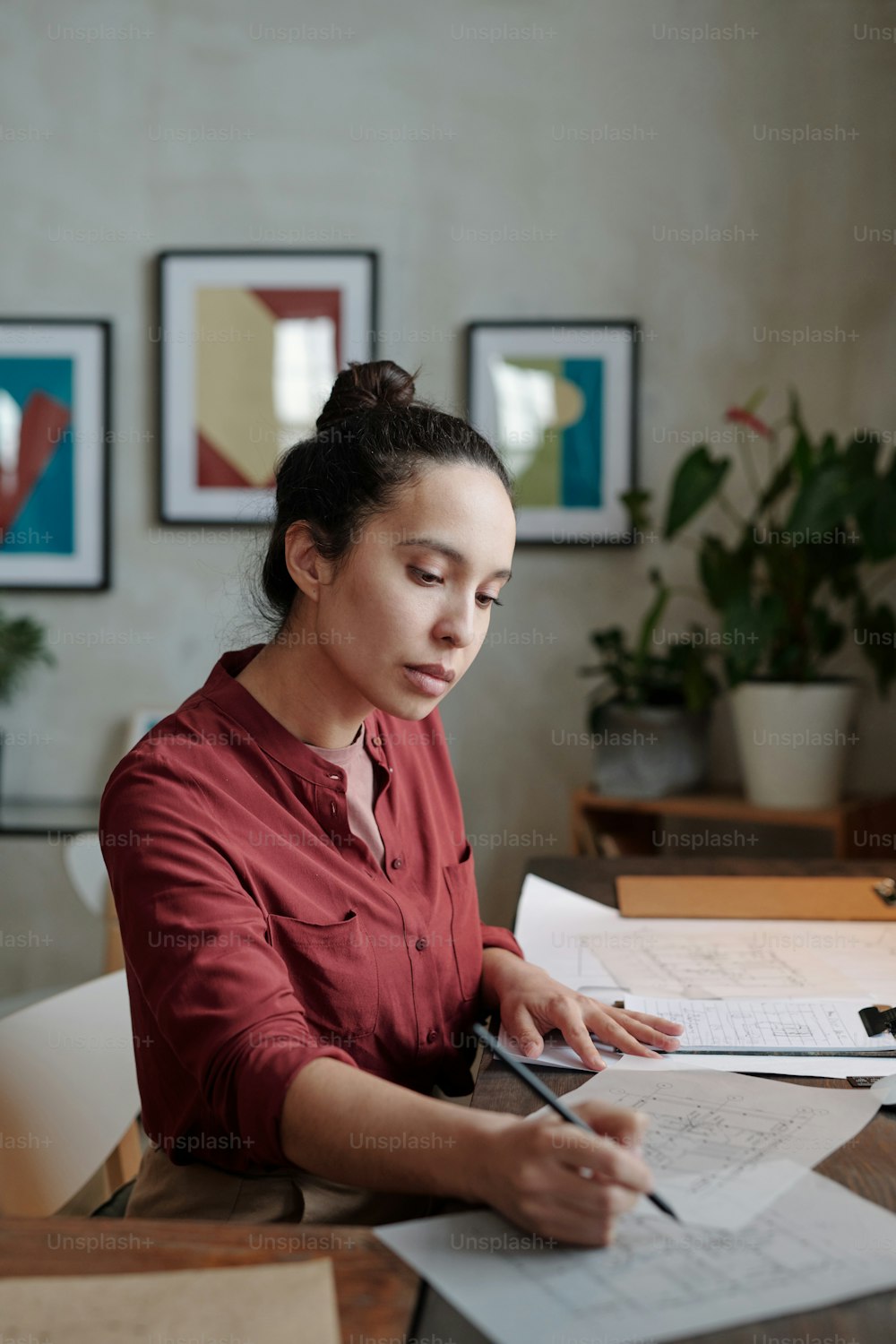 Mujer hispana joven concentrada en camisa burdeos sentada en el escritorio y dibujando bocetos mientras desarrollaba el diseño del edificio