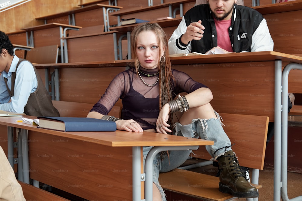 Bonita joven estudiante con ropa casual mirándote mientras está sentada junto a un largo escritorio de madera en la sala de conferencias entre compañeros de grupo masculinos interculturales