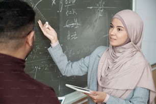 Young female student in hijab looking at professor while both standing by blackboard and girl pointing at graph while solving equation at lesson