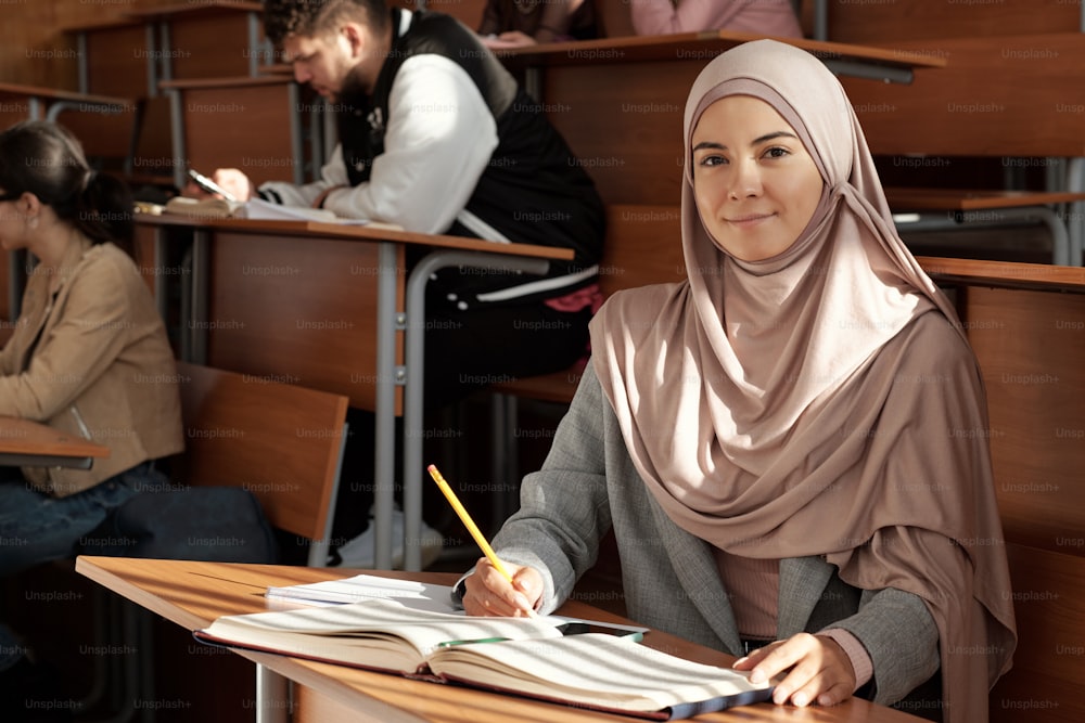 Young smiling female student in hijab sitting by desk at lesson in lecture hall and looking you while reading book against her groupmates