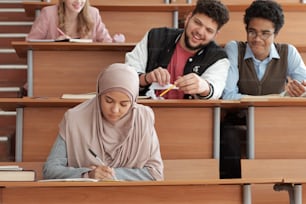 Menina séria em hijab fazendo anotações em copybook na palestra enquanto sentada à mesa contra dois colegas de grupo interculturais tirando sarro dela